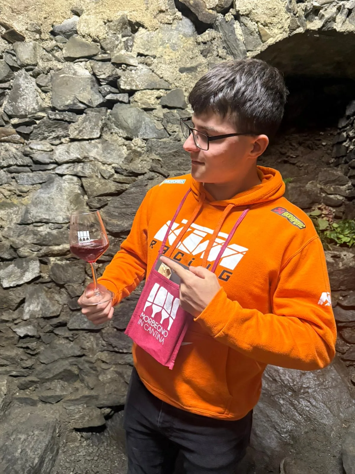Matteo Crespi Matteo con felpa KTM e un calice di vino in una cantina in pietra